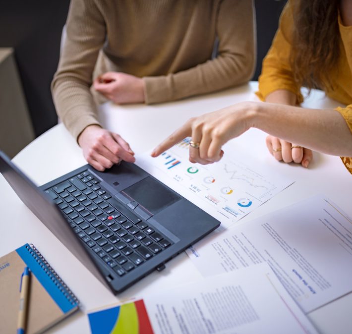 Two people sitting in front of a laptop at a small round table with documents on it, and one person pointing at the screen.
