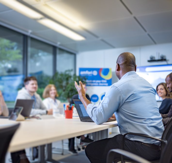Someone is talking in front of 7 people, all sitting around a table in a meeting room.