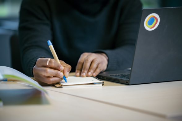 Someone sitting in front of a laptop and writing something in a notebook with a pen.