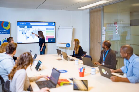 A meeting room with people sitting around the table with their laptops looking at someone standing next to a big screen and pointing at something on a world map.